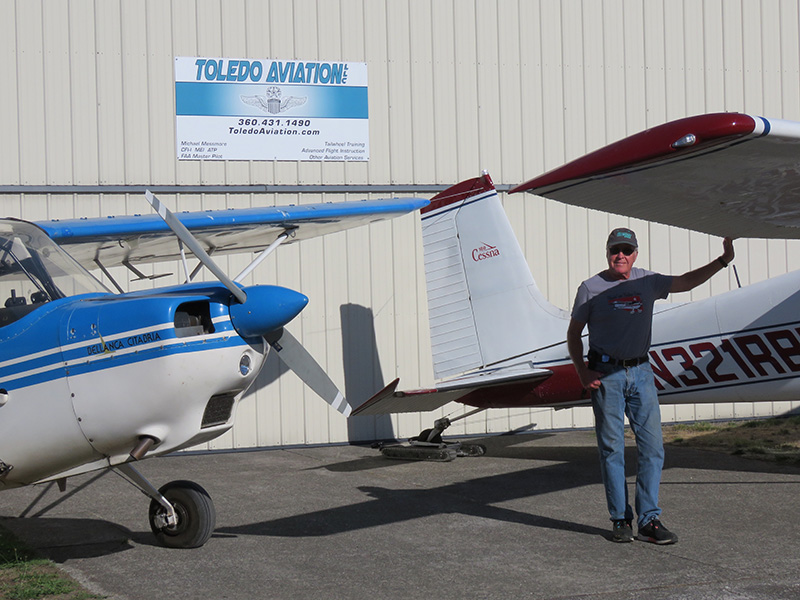 Pilot Michael Messmore stands between two small aircraft in front of a hangar at the airport.